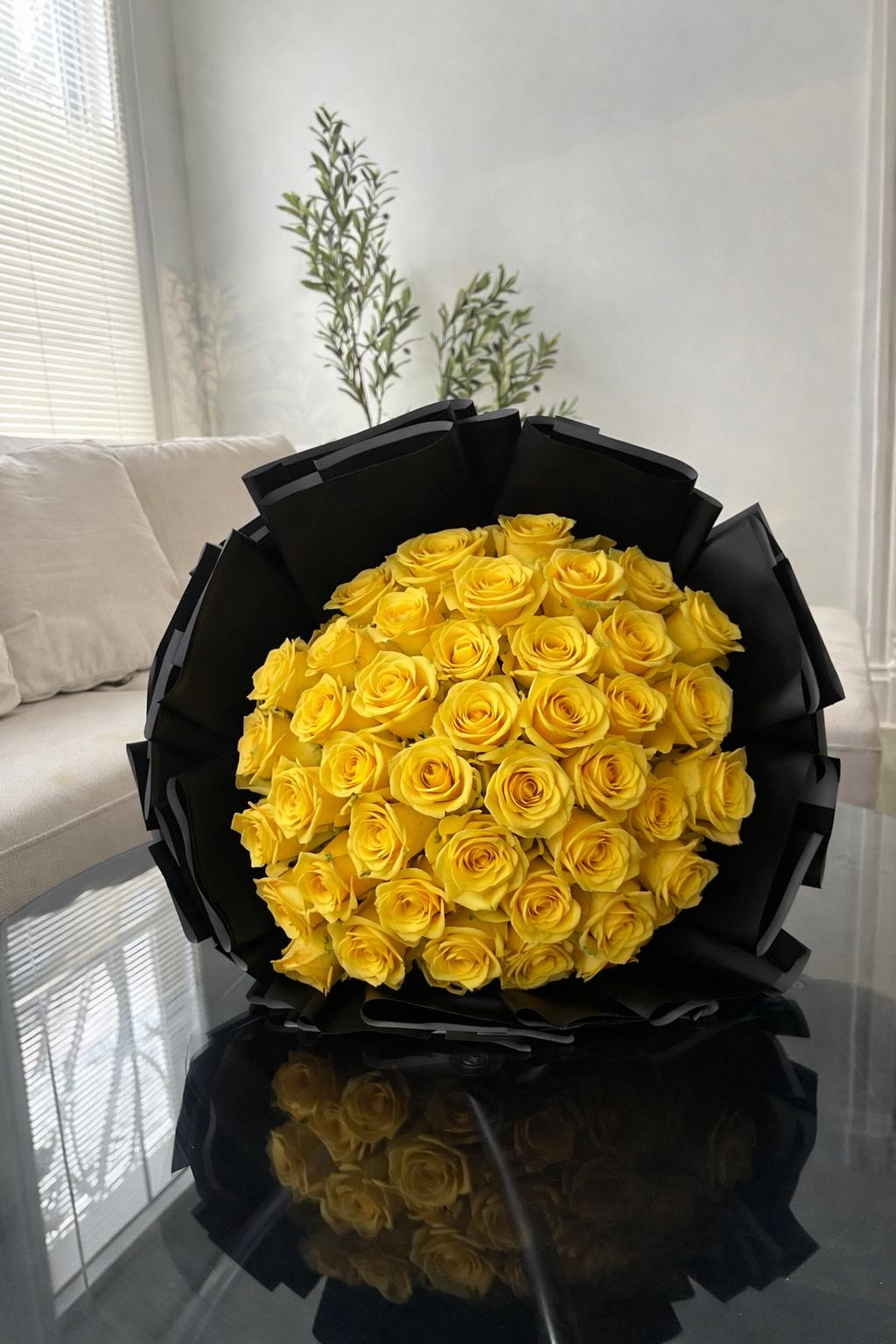 Bouquet of yellow roses in a black holder on a reflective surface with a light gray wall and white couch in the background.